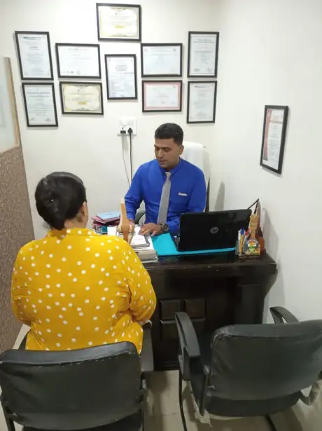 A patient seated across the desk from the physiotherapist during a consultation, with two rows of framed certificates on the wall behind them.
